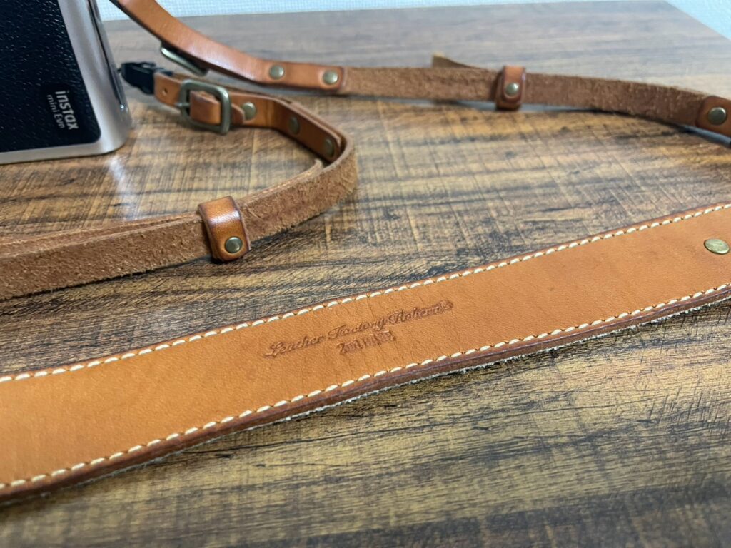 木目のテーブルに置かれた、私が選んだ「ROBERU」の本革カメラストラップ。ブランドロゴが刻印された深いブラウンのレザー。
