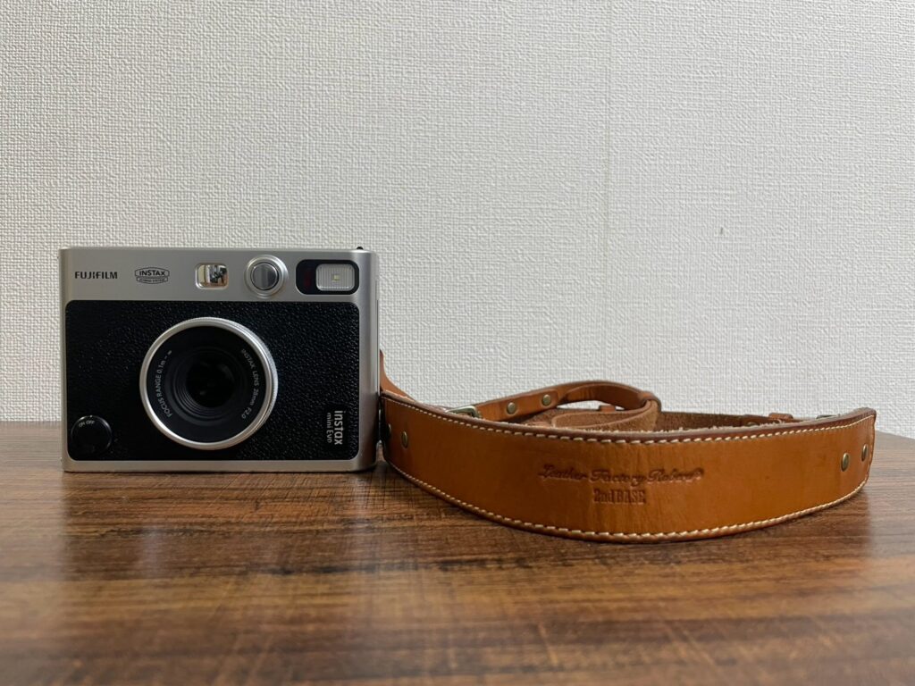 木目のテーブルに置かれたinstax mini Evoと「ROBERU」の本革カメラストラップ。クラシカルなカメラ本体と味わい深いレザーの組み合わせ。