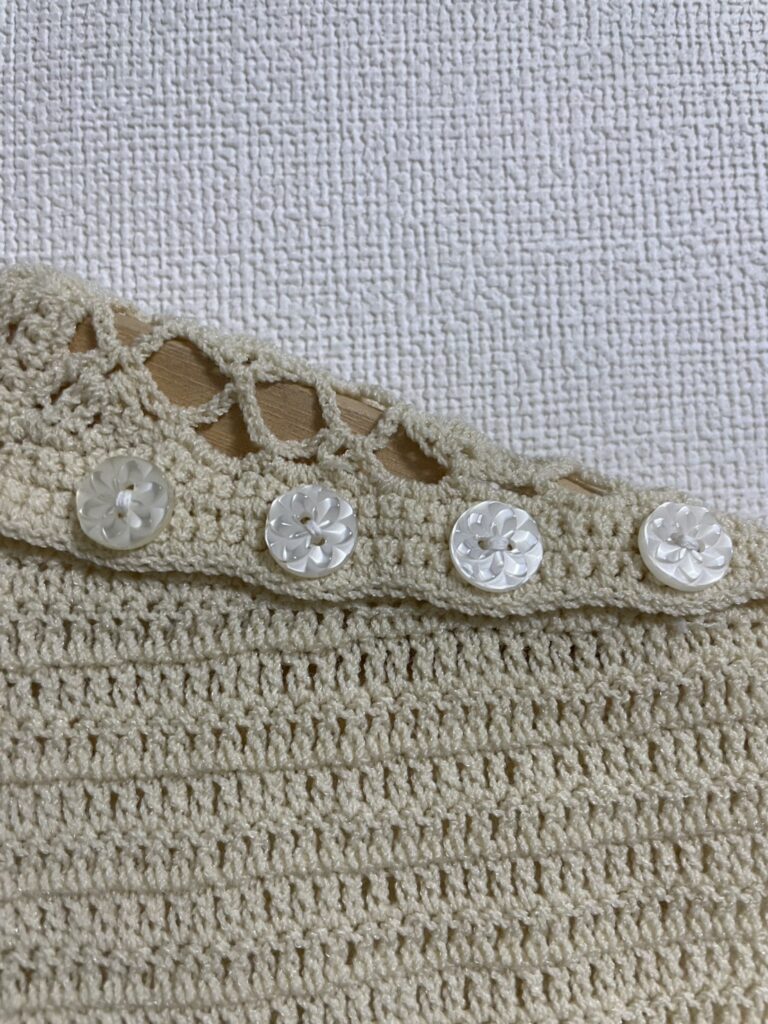 クリーム色の70sヴィンテージサマーニットにおける、小さなこだわり。見えるか見えないかわからない些細な部分だが、近接すると、厚みと透け感のある花柄のボタンが使用されていることがわかる。ニット全体と調和し、目立ちすぎないように配慮されたデザイン。